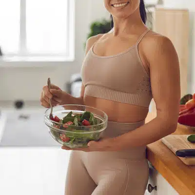 A smiling woman in fitness attire holds a bowl of salad in her bright kitchen