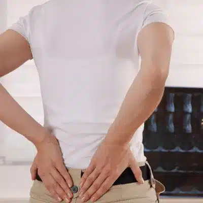 Person stands indoors, pressing lower back with both hands, experiencing localized lumbar ache today pain