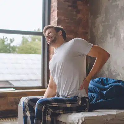 Man sitting on bed in plaid pants holding lower back, wincing from morning back pain stiffness