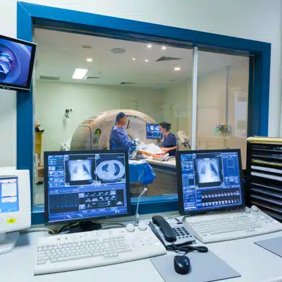 Technicians operate CT scanner from control room, monitoring patient positioning and images on multiple screens
