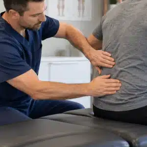A chiropractor gently examines a seated male patient's lower back using both hands to assess spinal mobility and determine how often should you go to the chiropractor for ongoing care